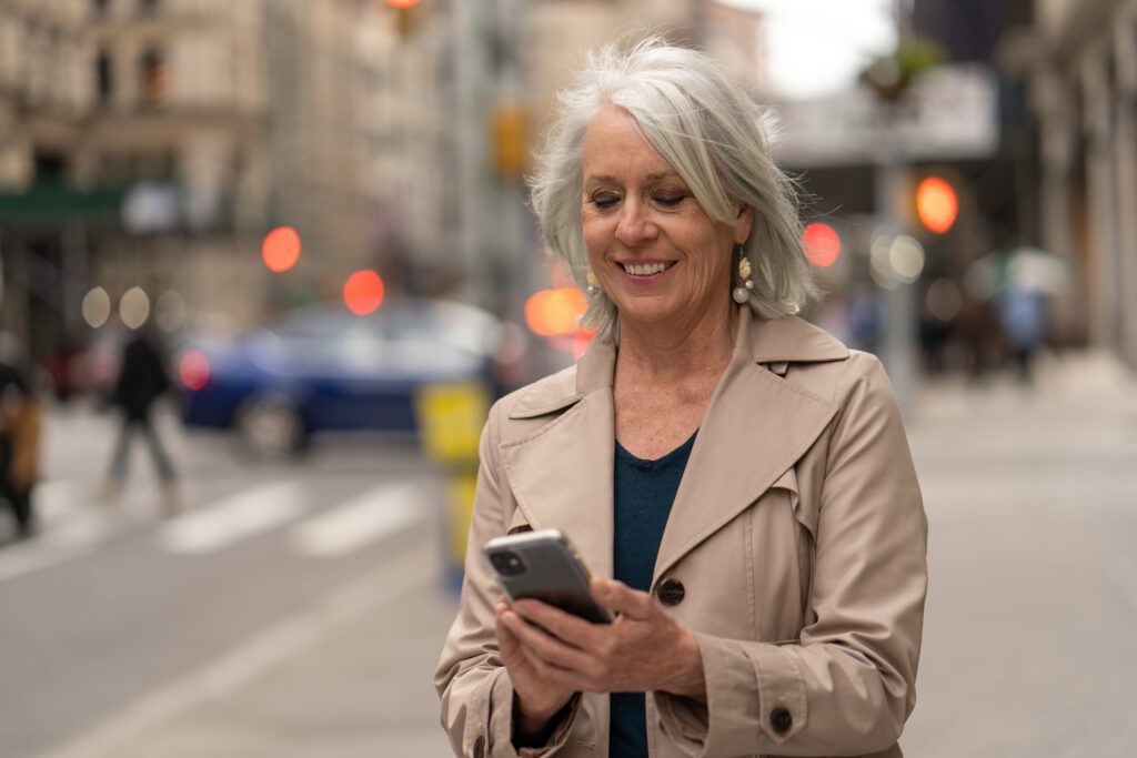 A mature white woman smiling and using her smartphone on the sidewalk of a busy street in New York City.