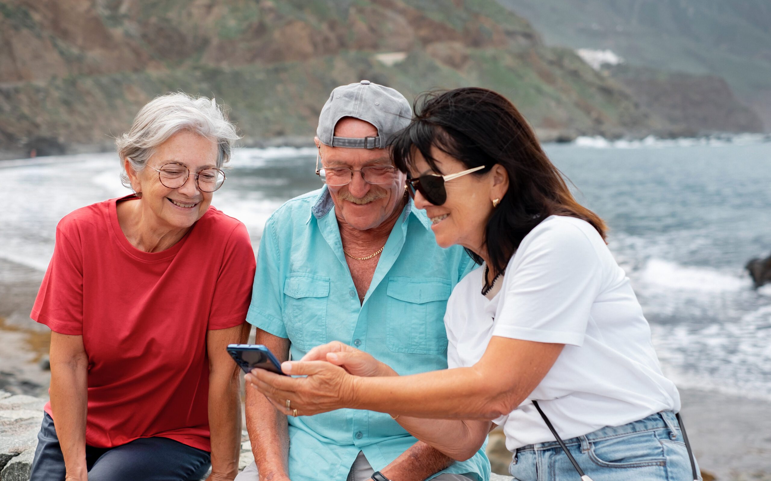 Happy elderly people sitting by the California shoreline using a smartphone. 