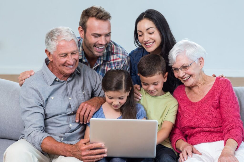 Happy multi-generational family household sitting together on a couch at home using a laptop. 
