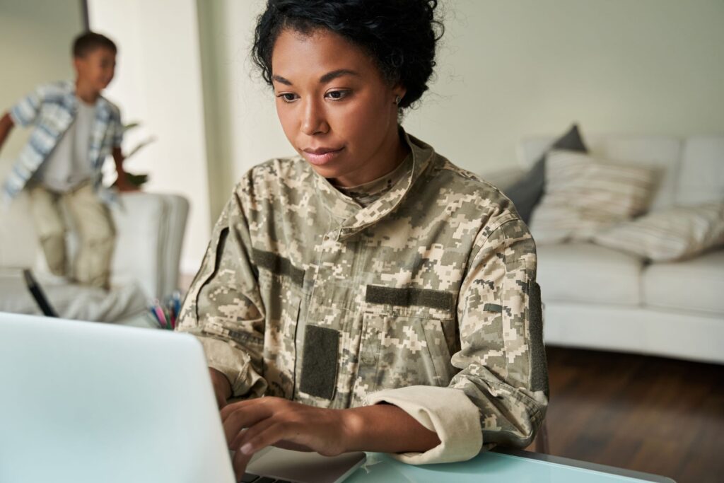 Front view of a young Black veteran woman in uniform using her laptop to apply for ACP online while her son plays in the living room behind her. 
