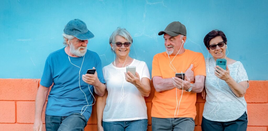Group of four seniors leaning against an orange and blue wall enjoying their free cell phone service and using their smartphones outside.