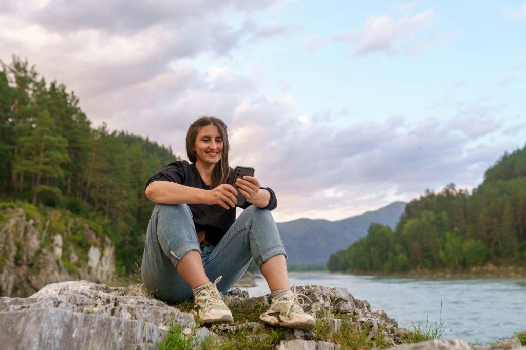Mature woman sitting alone on a rock outdoors near a river with forested woodlands in the background while happily using her cell phone.